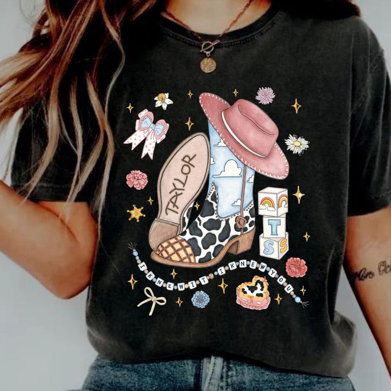Op de afbeelding: Een antracietgrijs t-shirt met een grafische print van een cowboylaars met koeienvlekken, een roze hoed, een strik en blokken met de letters T, S en een regenboog. De zool is gelabeld met TAYLOR en de tekst I KNEW YOU staat in een boog.