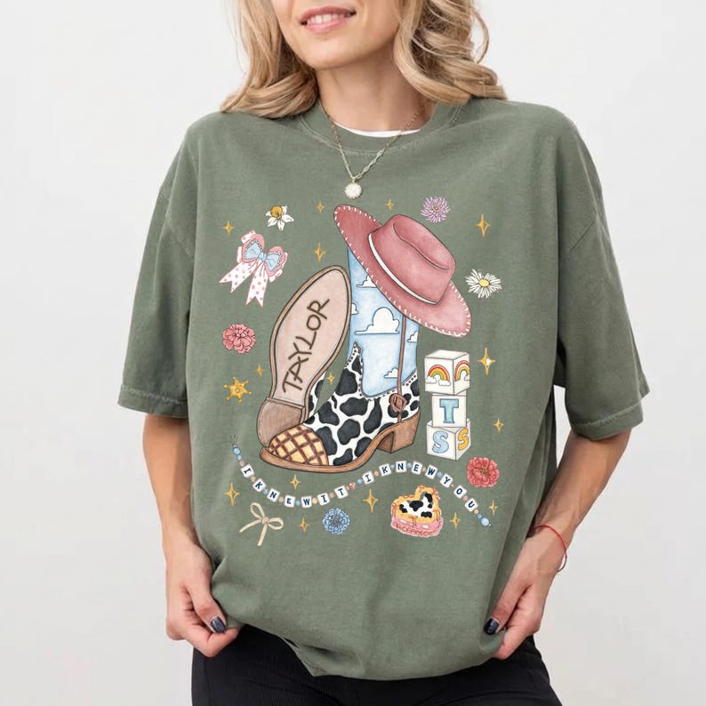 Op de afbeelding: Een olijfgroen T-shirt met korte mouwen en een grafische print van een cowboylaars met koeienprint, een roze hoed, een strik, bloemen en blokken met de letters T S. De tekst "TAYLOR" en "I KNEW IT I KNEW YOU" zijn op de print te zien.