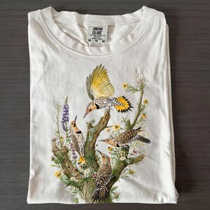 May include: An off-white short-sleeve t-shirt with a Comfort Colors brand label at the neck. The front features a detailed graphic of four northern flickers perched on and flying around a tree branch surrounded by colorful wildflowers and greenery.