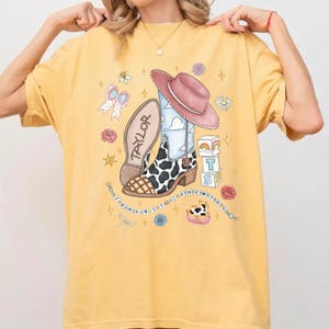 Op de afbeelding: Een mosterdgeel t-shirt met een grafische print van een cowboylaars met koeienprint, een roze cowboyhoed, een strik, bouwblokken met een regenboog en een kralensnoer met de tekst I KNEW YOU WERE TROUBLE en TAYLOR.