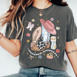 Op de afbeelding: Een antracietgrijs t-shirt met een grafische print van een cowboylaars met koeienvlekkenpatroon, een roze hoed, alfabetblokken met de letters TS, en de tekst TAYLOR en I KNEW YOU. Het ontwerp bevat bloemen, sterren en een strik.