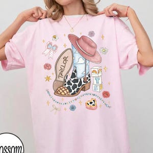 Op de afbeelding: Een lichtroze t-shirt met een grafische print van een cowboylaars met koeienprint, een roze cowboyhoed en een laarszool met het label Taylor. Het ontwerp bevat alfabetblokken, een kralensnoer met de tekst I KNEW IT I KNEW YOU en bloemen.
