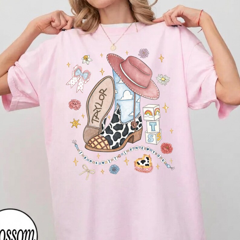 Op de afbeelding: Een lichtroze t-shirt met een grafische print van een cowboylaars met koeienprint, een roze cowboyhoed en een laarszool met het label Taylor. Het ontwerp bevat alfabetblokken, een kralensnoer met de tekst I KNEW IT I KNEW YOU en bloemen.