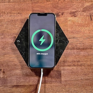 Puede incluir: Un smartphone dorado que muestra un símbolo de carga y el texto "95% Charged" en una base de carga de mármol negro y gris. El teléfono está conectado a un cable de carga blanco. El fondo es una superficie de madera.