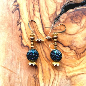 May include: A pair of handcrafted earrings featuring dark blue, iridescent glass beads with an ornate pattern. The earrings have gold-colored metal accents, including small beads, decorative caps, and ear wires. The background is a wooden surface.