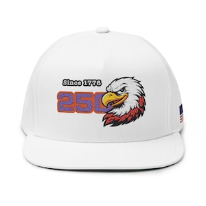 Peut inclure: Casquette de baseball blanche avec une tête d'aigle brodée en rouge, blanc et noir. La casquette porte le texte "Since 1776" au-dessus du chiffre "250" en violet et orange. Un petit drapeau américain est sur le côté.