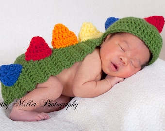 May include: A baby wearing a green, blue, red, yellow, and orange crocheted dinosaur costume. The baby is sleeping with its arms tucked under its chin.