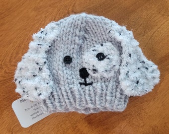 May include: Gray knitted dog hat with black button eyes and a white fuzzy nose. The hat has a tag that reads "Elbe, Size: Cozy & snuggly, 100% acrylic, Fiberfill".