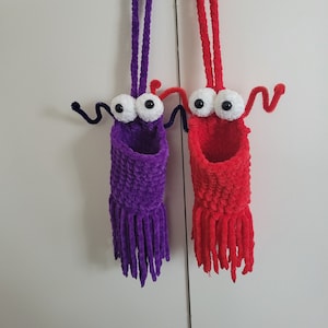 May include: Two handmade, whimsical wall organisers. One is purple, the other is red, both with large googly eyes, pipe cleaner arms, and fringe. They hang from white and black flower-shaped hooks.