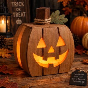 May include: A wooden jack-o'-lantern decoration with a carved face, illuminated from within by a warm light. The pumpkin features a square stem wrapped in twine, a single green fabric leaf, and vertical slats. A black tag reads: Handmade with Love for a Spooky Season!