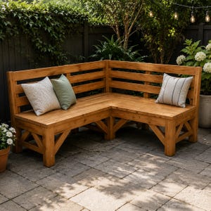 以下が含まれることがあります： L字型の木製コーナーベンチで、スラット状の背もたれとナチュラルな木目の仕上げが特徴です。ベンチには3つの装飾用クッションが置かれています。質感のある白い正方形のクッション、無地のセージグリーンのクッション、そして細い黒いストライプが入った白いクッションです。