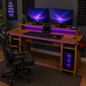May include: A wooden gaming desk with a hutch, two computer monitors displaying a purple nebula, a keyboard, a mouse, and a black gaming chair. The desk features integrated purple LED lighting, a cup holder, and a computer tower with glowing fans.