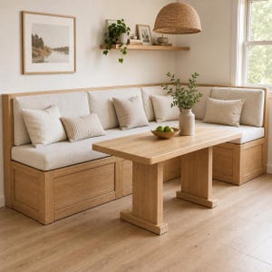 May include: A light oak corner bench with cream-colored upholstered cushions and matching throw pillows. A rectangular light oak dining table sits in front of the bench. A ceramic vase with green foliage and a bowl of green apples sit on the table.