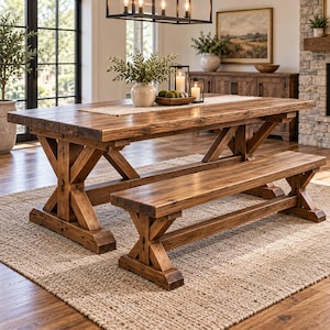 May include: A rectangular wooden dining table and a matching bench, both featuring a medium brown finish and sturdy X-shaped trestle legs. The table surface displays a natural wood grain texture, providing a rustic appearance for the dining set.