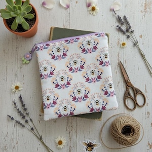 May include: A white zipped pouch featuring a bee and floral design, placed on a book. The pouch has a purple zip and a flower charm. The image also includes a succulent, flowers, scissors, and twine.