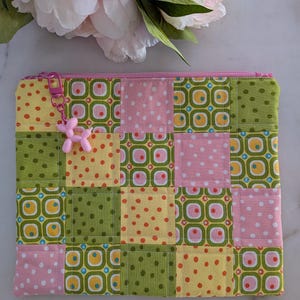 May include: A rectangular fabric zip pouch featuring a patchwork design of squares in shades of pink, yellow, and green with polka dot and geometric patterns. A small pink balloon dog charm is attached to the pink zip pull.