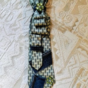 May include: A necktie featuring a light green fabric with a repeating dark geometric pattern, accented with dark blue denim fabric sections, a dark blue denim loop, and two decorative green flower-shaped embellishments with clear rhinestones.