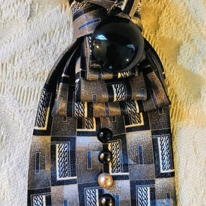 May include: A necktie featuring a geometric pattern in shades of gray, black, and blue. The tie is embellished with a large, round, glossy black button at the knot and a vertical row of alternating black and cream-colored beads down the center.