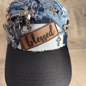 Puede incluir: Gorra de béisbol de mezclilla con visera negra. La gorra está adornada con varias cruces plateadas y doradas, pedrería y un parche marrón que dice "blessed". La mezclilla tiene un aspecto desgastado.