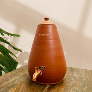 May include: A terracotta-colored clay water dispenser with a spigot and lid. The dispenser has a rounded, teardrop shape and the text "RustiClay" is visible on the front. The spigot is made of a copper-colored metal.