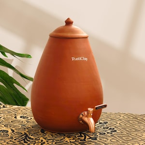 Puede incluir: Un dispensador de agua de arcilla de terracota con un grifo de color cobre. El dispensador tiene un cuerpo redondeado, una tapa y el texto "RustiClay" en la parte delantera. El dispensador está sobre una superficie estampada.