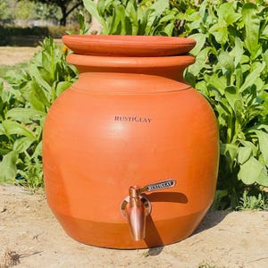 Puede incluir: Un recipiente de agua grande y redondo de terracota con tapa. El recipiente tiene un grifo y el texto "RUSTICLAY" es visible en la parte delantera. El recipiente de agua está sobre un fondo de follaje verde.