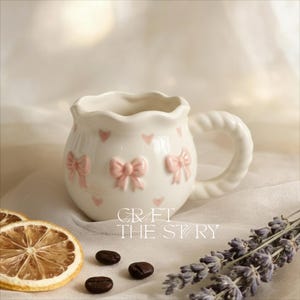 May include: A white ceramic mug with a scalloped rim and a twisted handle. The mug is decorated with pink bows and hearts. The text "CRAFT THE STORY" is printed on the mug.