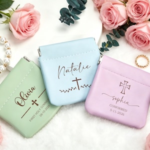 Puede incluir: Tres pequeñas bolsas personalizadas en colores pastel: verde menta, azul claro y lavanda. Cada bolsa presenta un nombre, una cruz y una fecha especial. Una pulsera de perlas y rosas rosas rodean las bolsas.