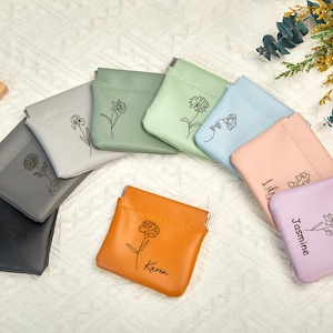 Può includere: Una collezione di piccole pochette colorate in varie tonalità, tra cui grigio, verde, blu e arancione. Ogni pochette presenta un motivo floreale e un nome, come "Karen" e "Jasmine". Le pochette sono disposte su una superficie bianca.
