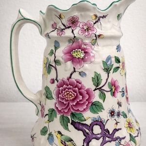 May include: A white ceramic pitcher with a floral design. The pitcher features a large pink peony, smaller pink and yellow flowers, and a colorful bird. The handle and rim are outlined in green.