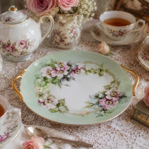 Könnte beinhalten: Vintage Teeservice mit hellgrüner Servierplatte mit floralem Design mit rosa und weißen Blumen. Das Set beinhaltet eine Teekanne, eine Zuckerdose, Teetassen und einen Teller mit Makronen. Goldakzente sind sichtbar.