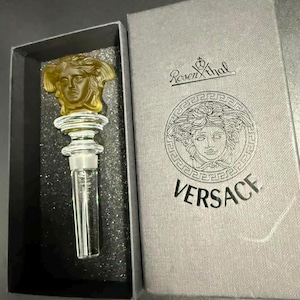 May include: A clear glass bottle stopper featuring a golden Medusa head topper, resting in a dark gray box. The box lid displays the Rosenthal logo and the Versace name alongside a circular Medusa head graphic printed in a dark gray ink.