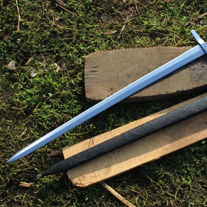 May include: A silver sword with a cross-shaped hilt and a brown and silver handle rests on a wooden plank. A black scabbard lies beside it. The sword is a medieval-style weapon.