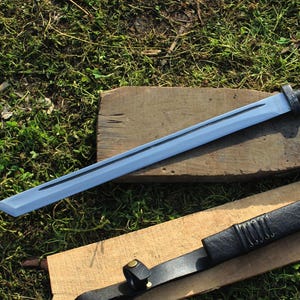 以下が含まれることがあります： 木製の板の上に、水色の刃と黒い柄の刀が置かれています。革のストラップが付いた黒い鞘が下にあります。この刀は武道やコスプレ用に設計されています。
