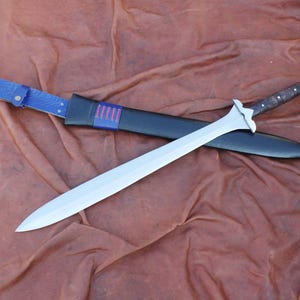 May include: A silver sword with a curved blade and a brown handle rests on a black sheath. The sheath is placed on a brown surface. The sword's handle has a silver pommel. The sheath has a blue strap.
