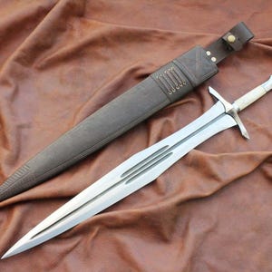 Puede incluir: Una espada plateada con una empuñadura en forma de cruz y un mango de color claro descansa junto a una vaina de cuero marrón. La vaina es de color marrón oscuro con una correa de cuero y cierre de botón. La hoja de la espada tiene tres ranuras paralelas.