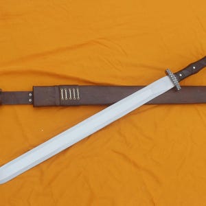 May include: A silver sword with a brown leather handle and sheath. The sword has a straight blade and a decorative hilt. The sheath is brown with a belt loop and a buckle. The background is a solid orange colour.