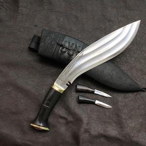 May include: A large, curved blade knife with a silver finish, black handle, and two smaller knives. The knife rests on a dark surface next to a black sheath. The blade has a decorative etched design.