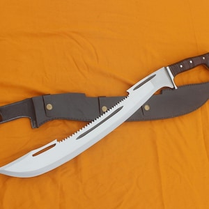 Puede incluir: Una gran machete plateado con borde dentado y mango marrón descansa sobre una funda de cuero marrón. La hoja tiene un diseño curvo con punta. El mango está envuelto en material marrón.