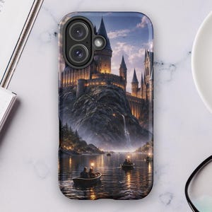 Puede incluir: Una funda de teléfono con una ilustración detallada de un castillo en una montaña, con un lago y botes en primer plano. La funda tiene un acabado brillante. La imagen incluye una taza de café, gafas y un cuaderno.