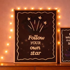 Könnte beinhalten: Gerahmte schwarze Tafel mit den weißen Worten "Follow your own star". Das Kunstwerk zeigt Sternschnuppen und Sterne. Der Rahmen ist hellbraun. Dekorative Lichter sind um den Rahmen herum angebracht.