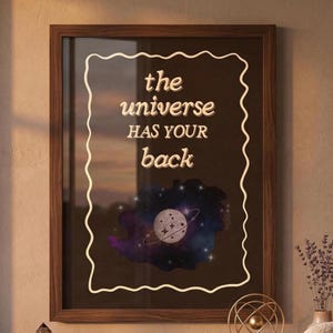 Könnte beinhalten: Gerahmter Kunstdruck mit dem Text "the universe HAS YOUR back" in einem weißen, wellenförmigen Rahmen. Das Kunstwerk zeigt ein himmlisches Design mit einem Planeten und Ringen, vor einem dunklen Hintergrund mit lila und blauen Farbtönen. Der Rahmen ist dunkelbraun.