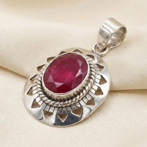 May include: A silver pendant with an oval, faceted, deep red gemstone. The gemstone is set in a decorative silver frame with a rope-like border and openwork design. The pendant has a loop for a chain.