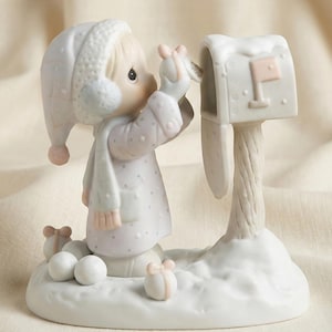 Puede incluir: Una figura de porcelana representa a un niño con ropa de invierno colocando un regalo en un buzón. El niño lleva un gorro, bufanda y abrigo blancos con detalles en rosa. El buzón blanco tiene una bandera rosa y la base presenta nieve y pequeños regalos.