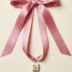 May include: A pink satin ribbon bow with long, flowing tails. A small, square pendant with a swan design hangs from the ribbon. The pendant is cream-colored with black detailing. The background is a neutral beige.