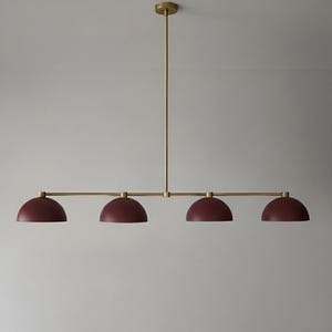 Op de afbeelding: Een moderne hanglamp met vier bordeauxkleurige, koepelvormige kappen. De armatuur heeft een messing afwerking en wordt aan het plafond gehangen door een centrale stang en horizontale armen.