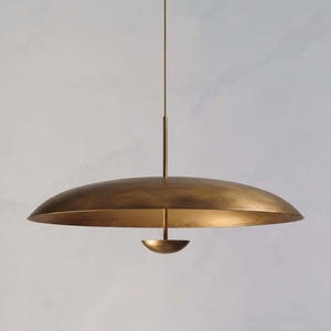 May include: A brass-colored pendant light fixture with a large, inverted saucer-shaped shade. A smaller, bowl-shaped element hangs below the shade. The fixture is suspended from a thin rod. The overall design is minimalist and modern.