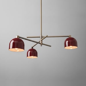 Op de afbeelding: Een moderne kroonluchter met vier bordeauxrode koepelvormige lampenkappen. De lamp heeft een messingkleurige centrale stang en armen, met de kappen gelijkmatig verdeeld. Elke kap heeft een gloeilamp.