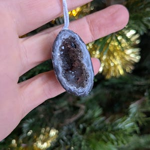 May include: An oval geode ornament suspended by a light blue string. The geode features a grey and white outer layer, enclosing a dark brown and crystal-filled core. The ornament is held in a hand, set against a blurred backdrop of green Christmas tree foliage.
