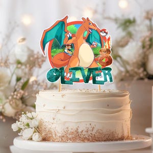 May include: A white frosted cake with a decorative topper featuring a cartoon Charizard character, a Pokeball, and the name "OLIVER" in teal letters. The cake is decorated with gold sprinkles and white flowers.